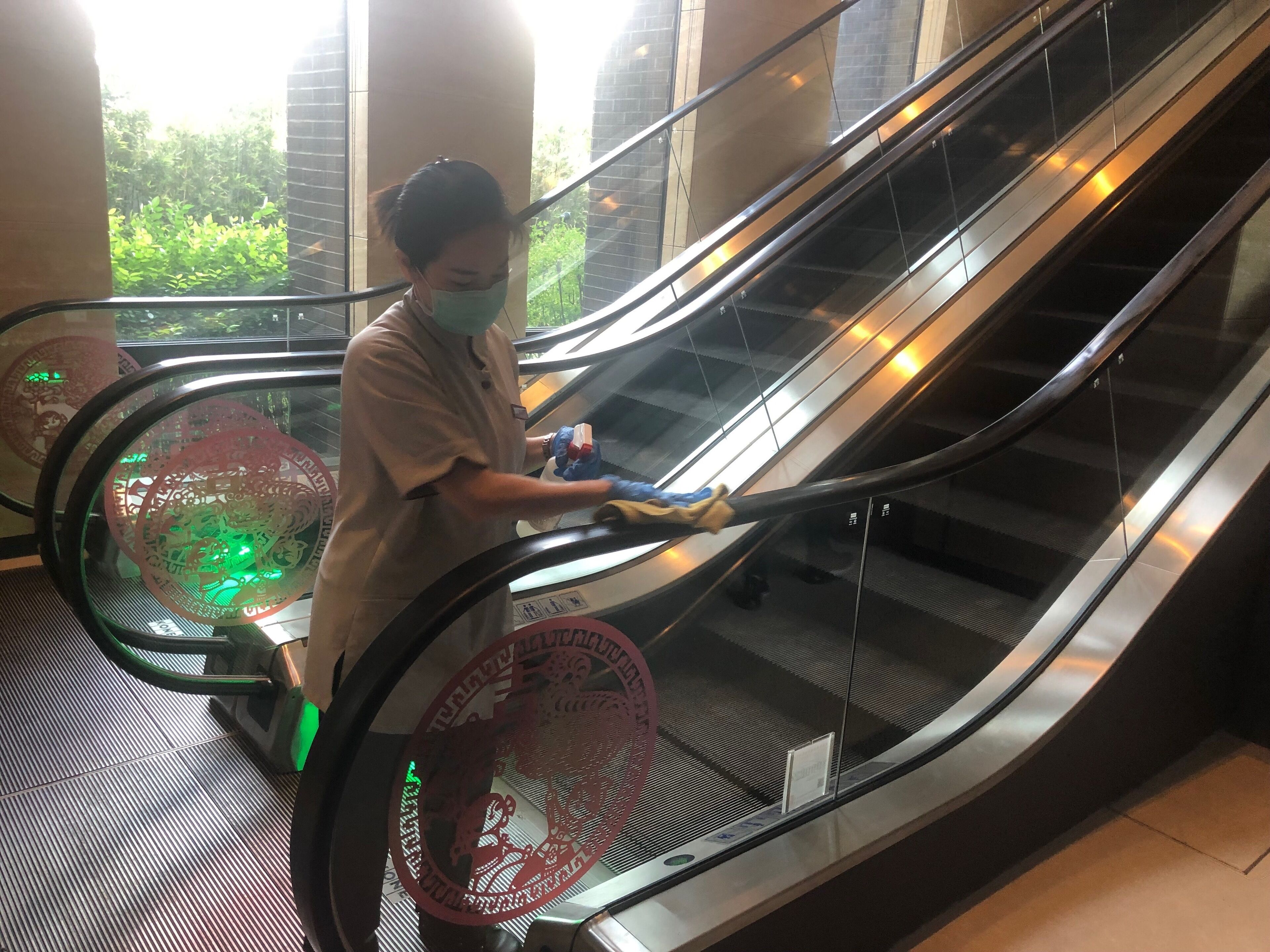 An escalator with a person standing on it, wearing a mask and gloves, holding a cleaning cloth.