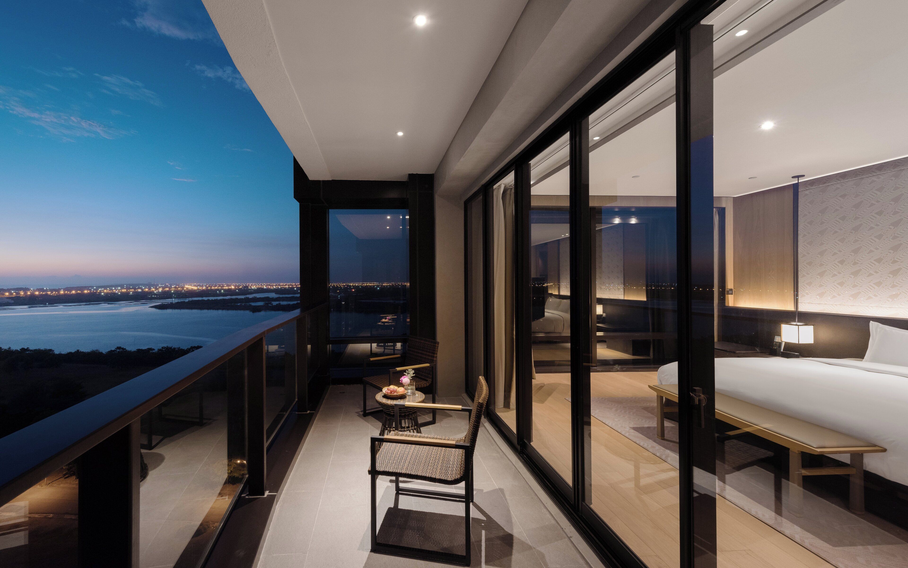 A modern hotel room with a balcony, a bed, a bedside table, and a view of the cityscape at dusk.
