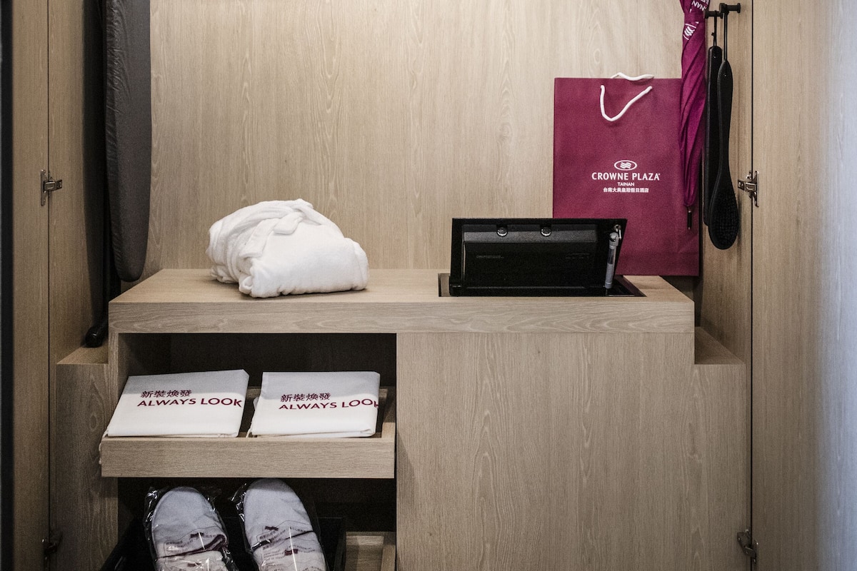 A hotel room with a wooden wardrobe, a shoe rack, a pair of shoes, a white towel, a black suitcase, and a red bag.