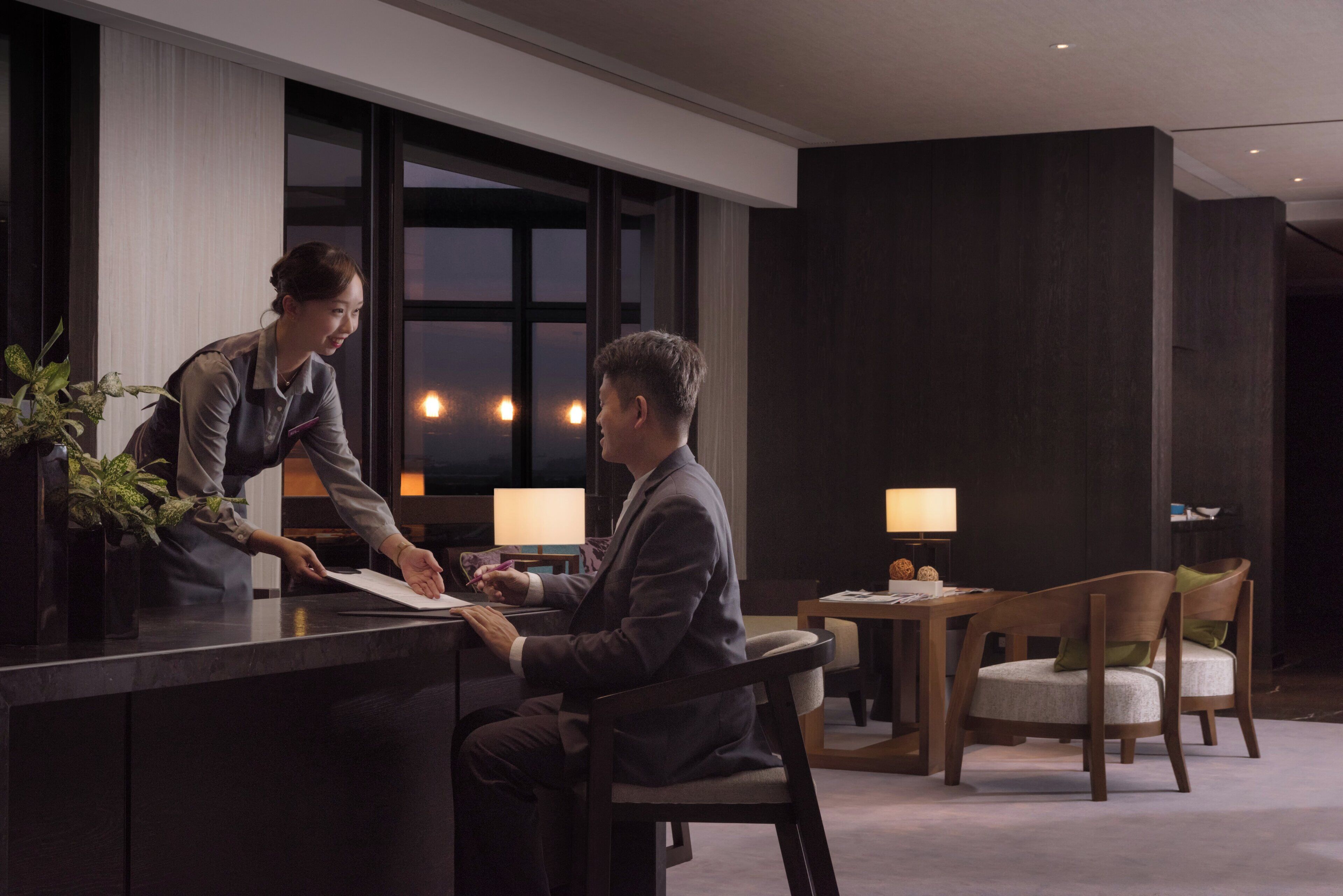 Zona de recepción de un hotel con un mostrador, un huésped sentado y un miembro del personal de pie. Hay dos lámparas sobre una mesa al fondo.