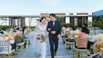 Una ceremonia de boda con invitados sentados en sillas doradas, una pareja caminando por un pasillo y un edificio al fondo.