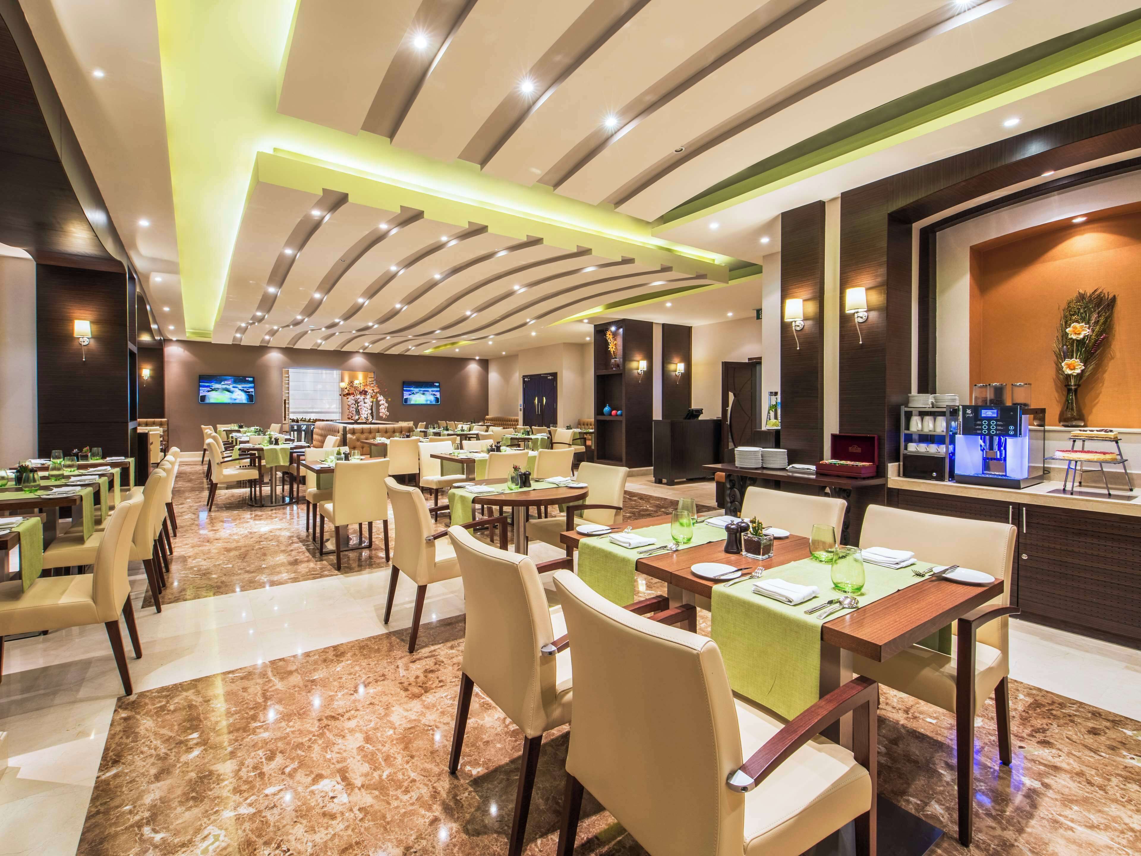 Un comedor de hotel espacioso con numerosas mesas y sillas, una zona de bar central y una pantalla de televisión montada en la pared.