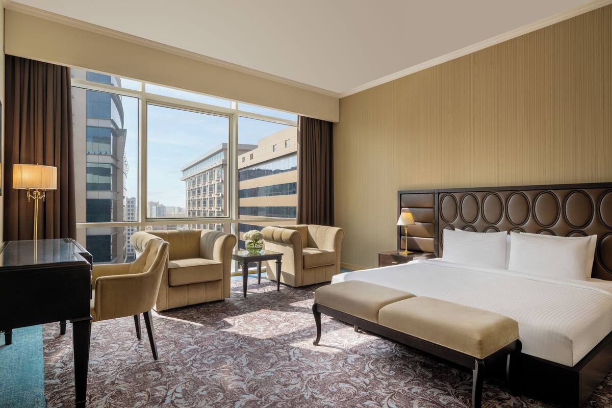 Une chambre d’hôtel avec un grand lit, deux fauteuils, une petite table avec un vase et un bureau avec une lampe.