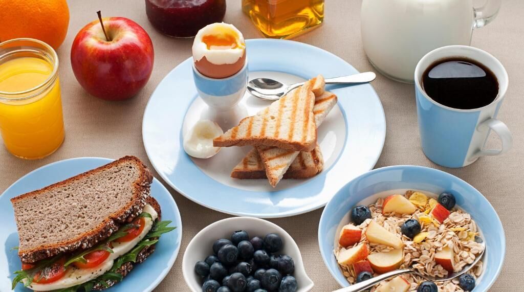 Un desayuno con tostadas, un tazón de arándanos, un plato de cereal con fruta, un vaso de jugo de naranja, una manzana, un frasco de mermelada, una taza de café y una tetera.