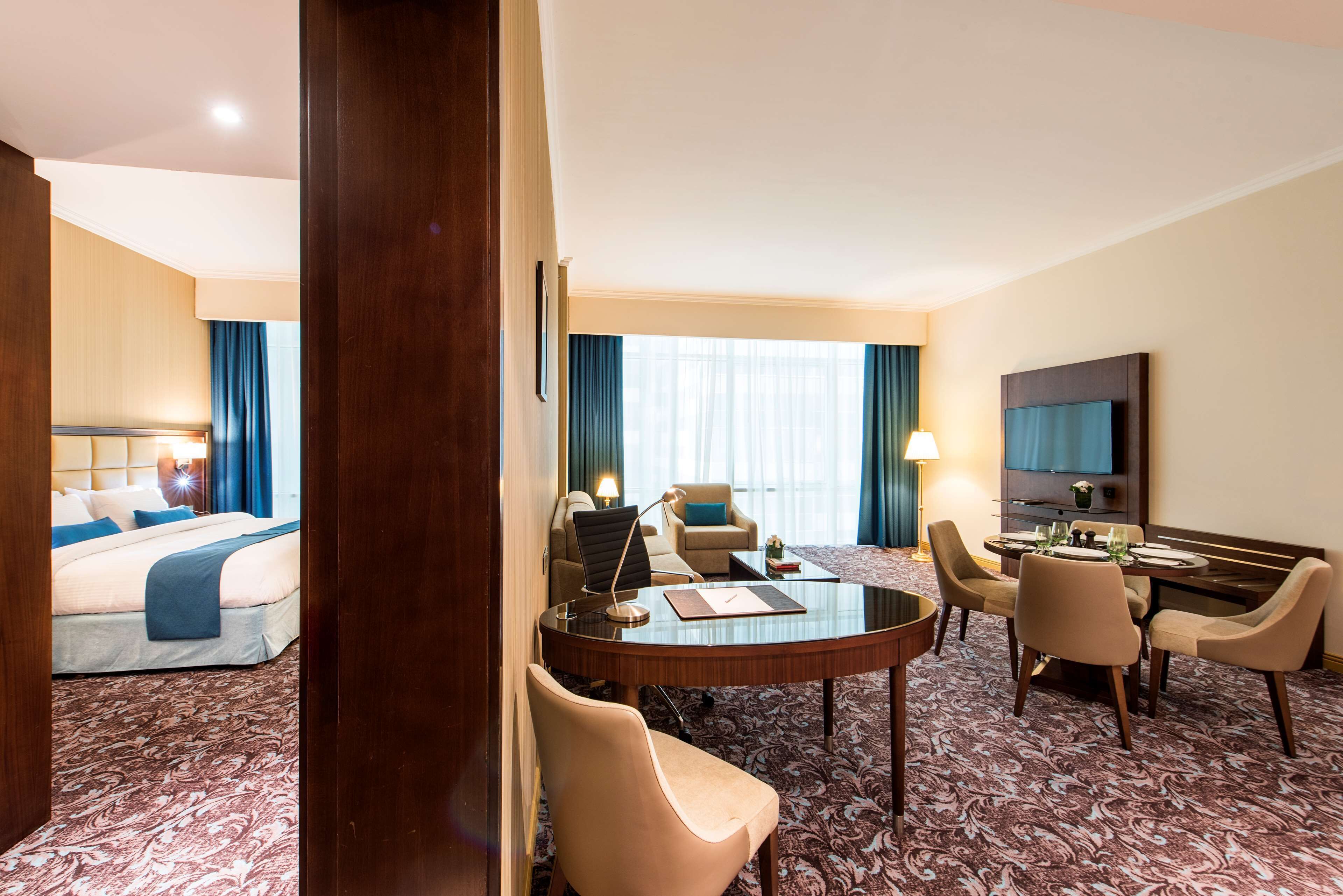 A hotel room with a bed, a desk, a dining table with chairs, a TV, and a large window with curtains.