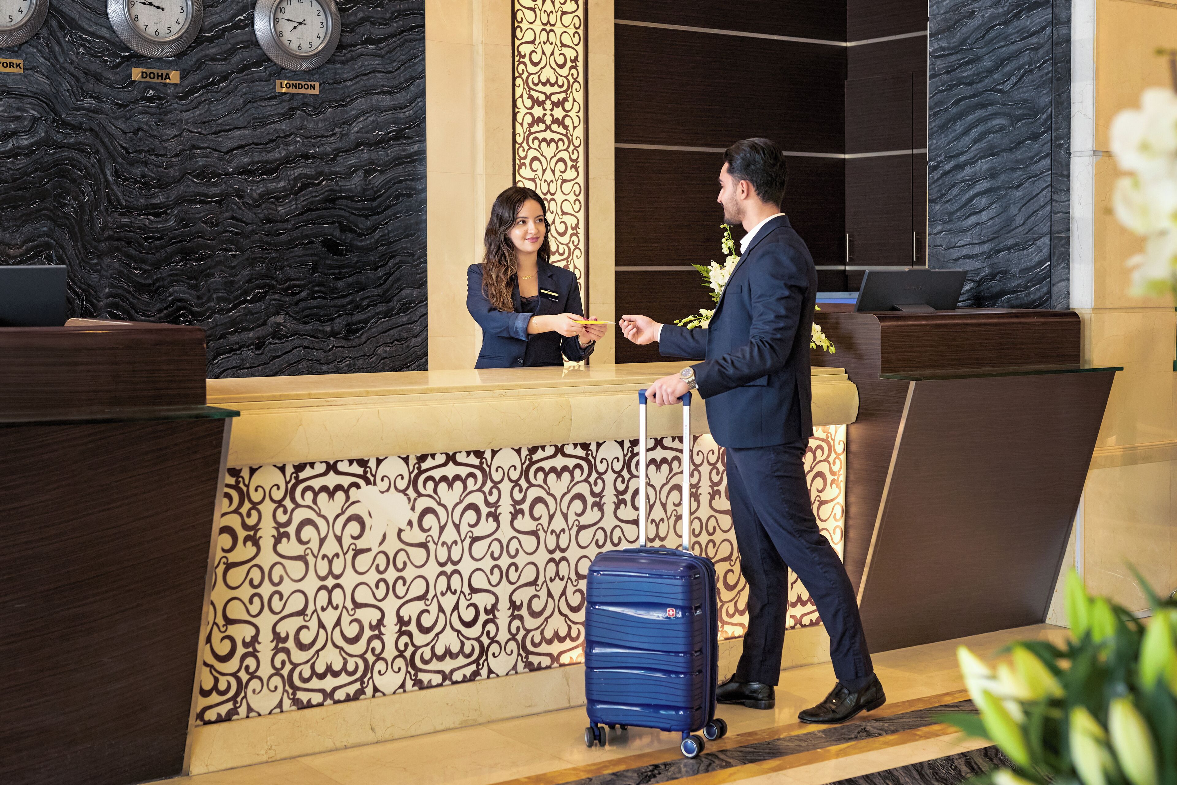 A hotel reception area with a receptionist, a guest, and a blue suitcase.