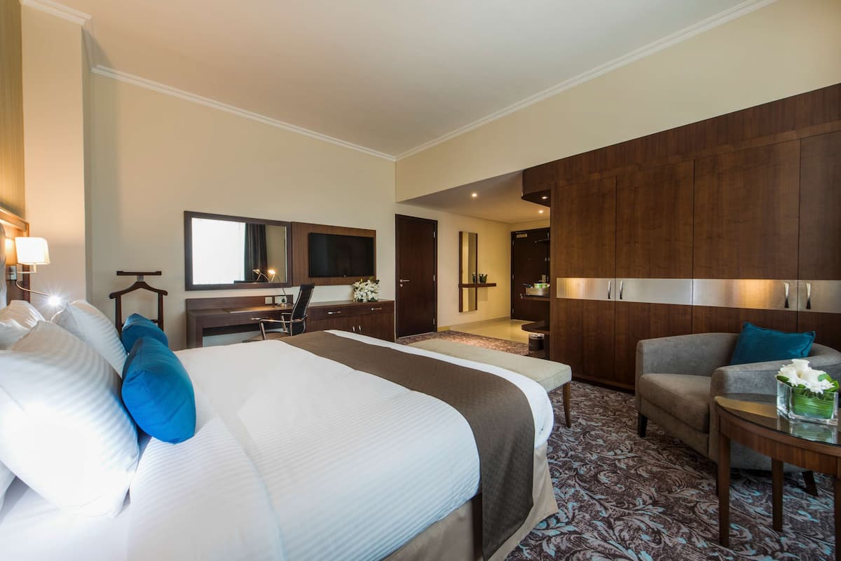 Une chambre d’hôtel avec un grand lit, un bureau, une chaise, un canapé et une salle de bain visible à travers une porte ouverte.