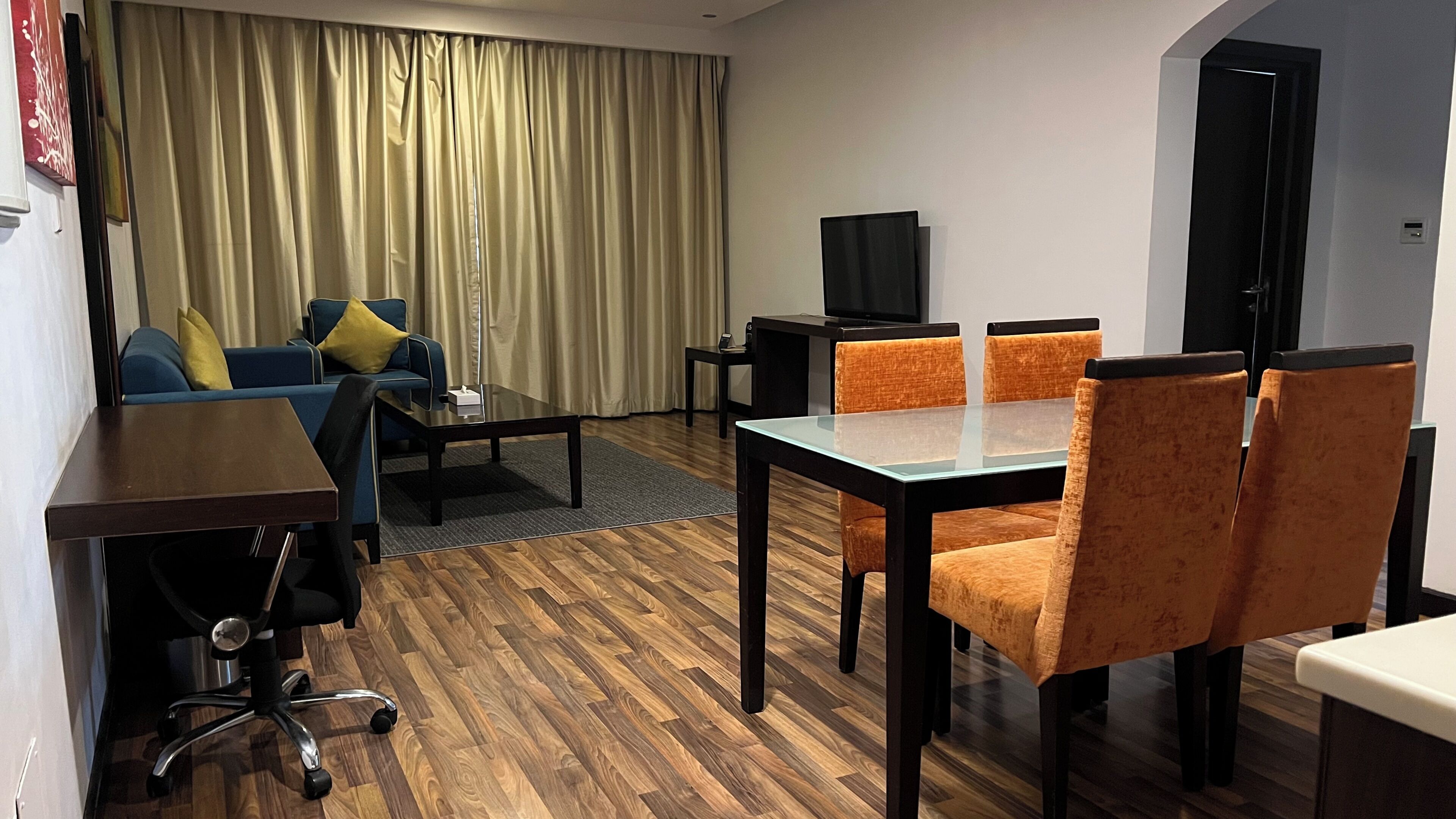 A hotel room with a sofa, a dining table with chairs, a TV, and a wooden floor.