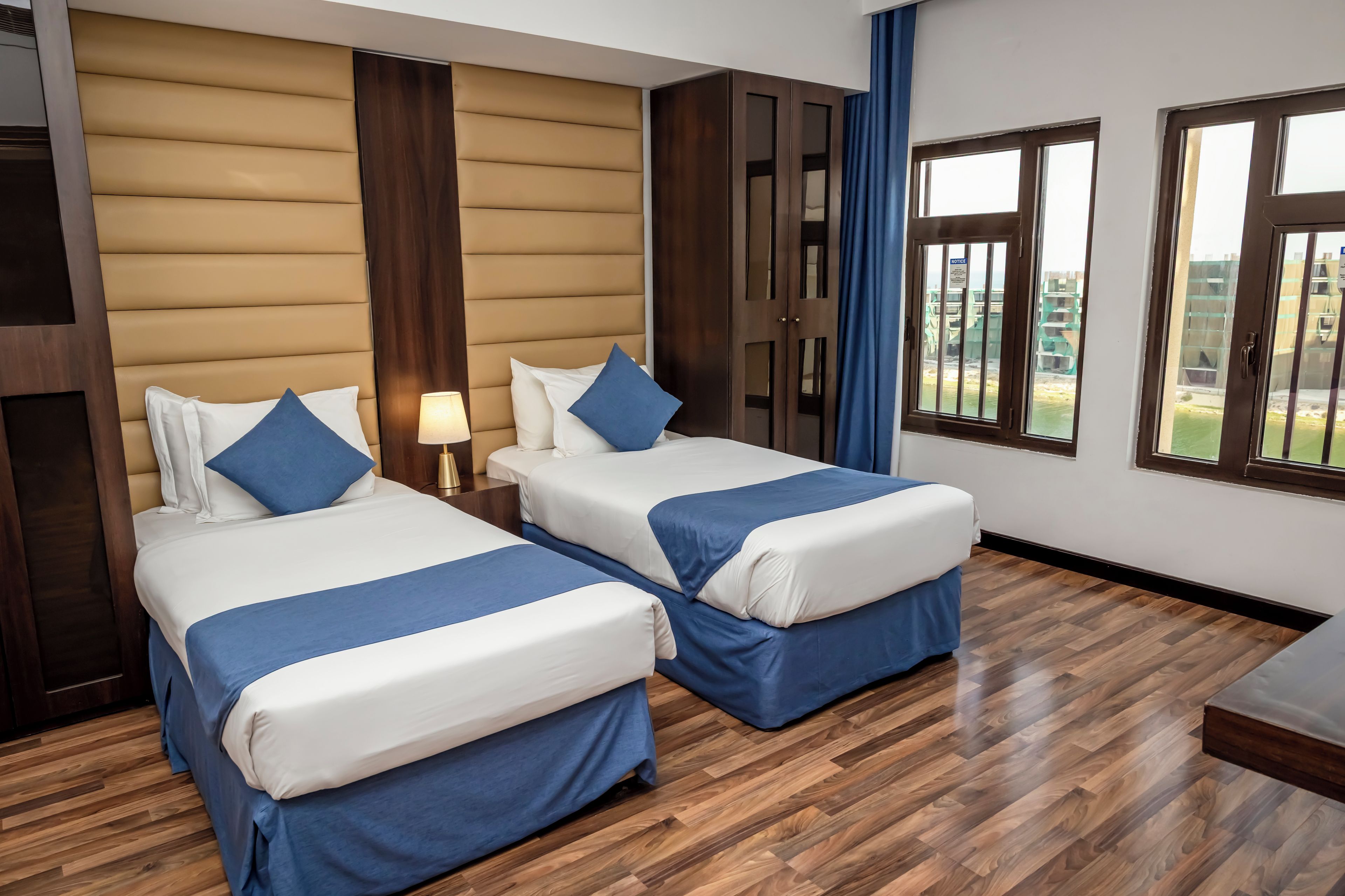 A hotel room with two beds, wooden flooring, a large window, and a view of the outside.