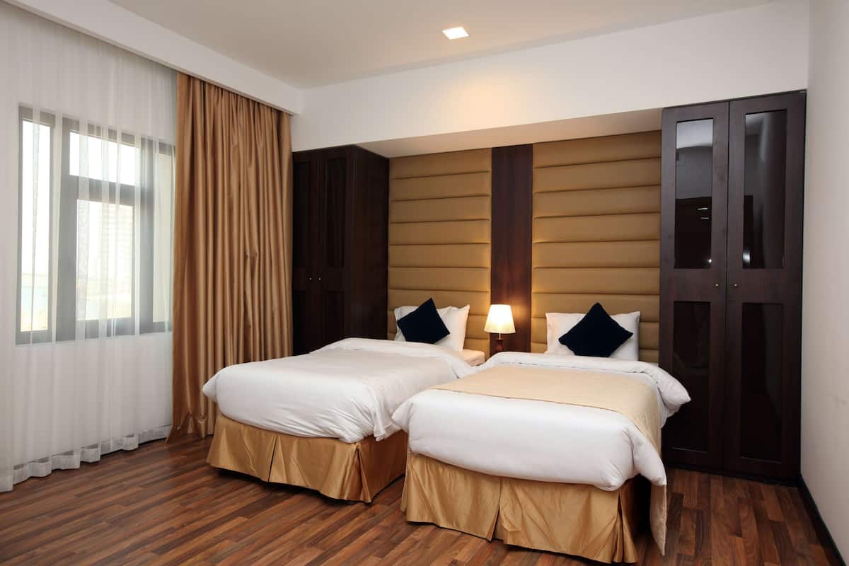 Ein Hotelzimmer mit zwei Betten, Holzboden, großem Fenster mit Vorhängen, dunklem Holzschrank und einer Wandlampe.