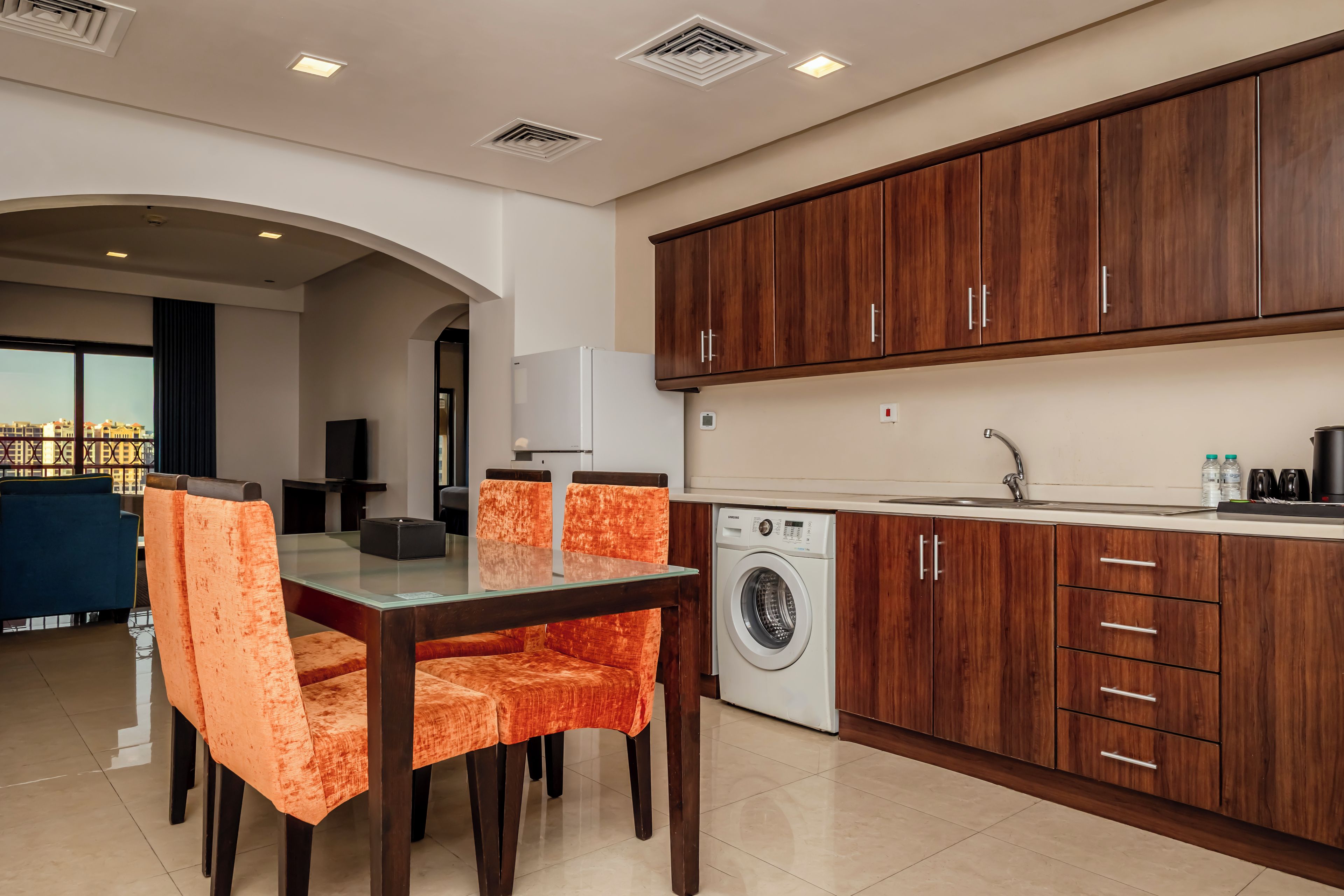 A modern kitchen with wooden cabinets, a built-in washing machine, and a dining area with orange chairs.