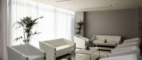 A modern, minimalist living room with white furniture, large windows with sheer curtains, and potted plants.