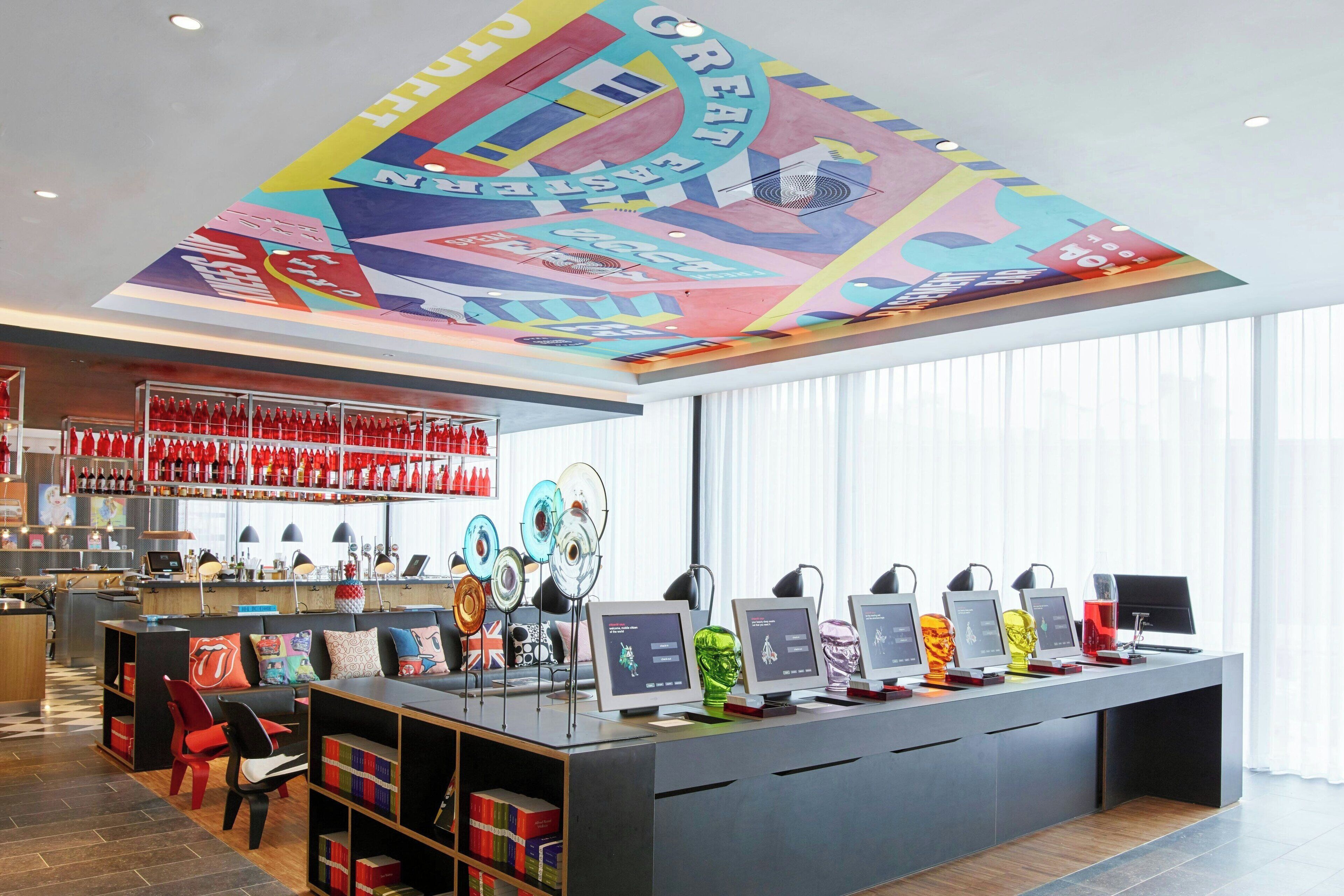 A modern bar area with a colorful ceiling mural, a long counter with multiple monitors, and a seating area with red chairs and bookshelves.