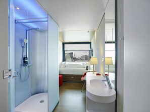 A modern hotel bathroom with a glass-enclosed shower, a white bathtub, a large window with a city view, and a separate sink area with two lamps. - citizenM London Shoreditch (London)