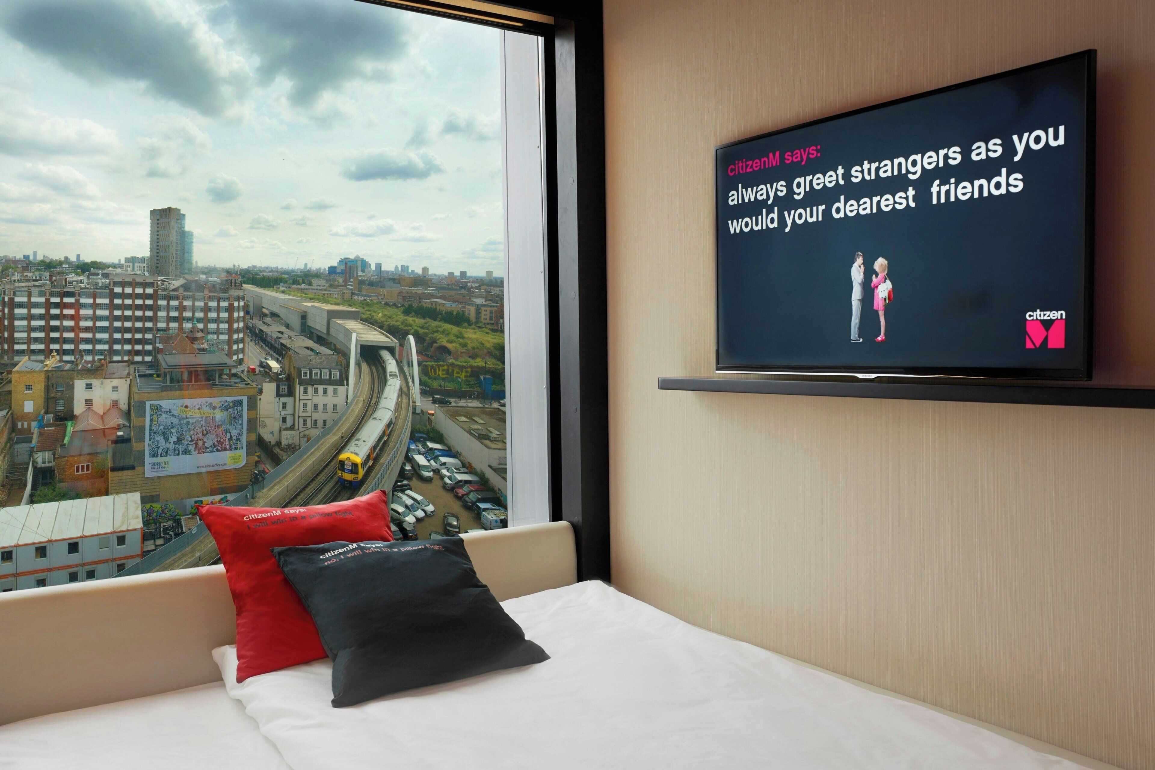 A hotel room with a bed, a television displaying a message, and a view of a cityscape through a window.