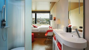 A modern hotel room with a shower, a bed with red accents, a white sink, and a large window offering a city view. - citizenM London Shoreditch (London)