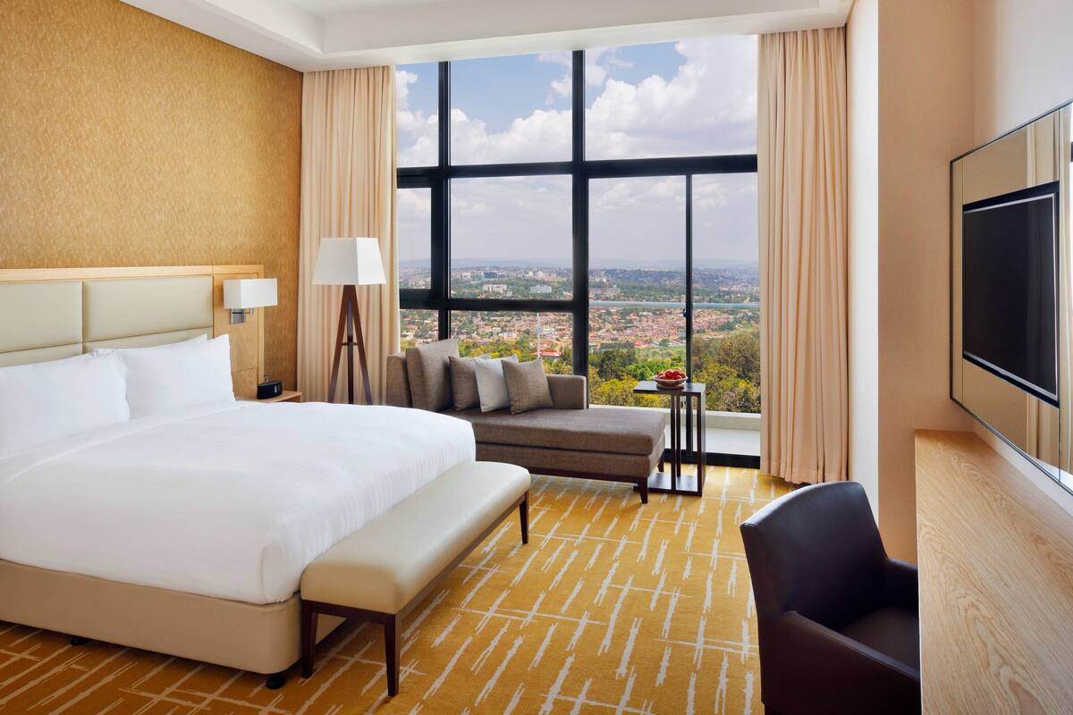 A hotel room with a large bed, a sofa, a TV, and a view of a cityscape through large windows.