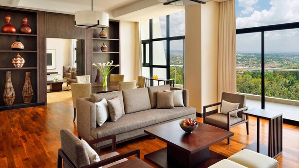 Amplia sala de estar con un sofá grande, sillones, una mesa de centro y un comedor. Hay una estantería con objetos decorativos y vistas a un paisaje urbano a través de amplias ventanas.