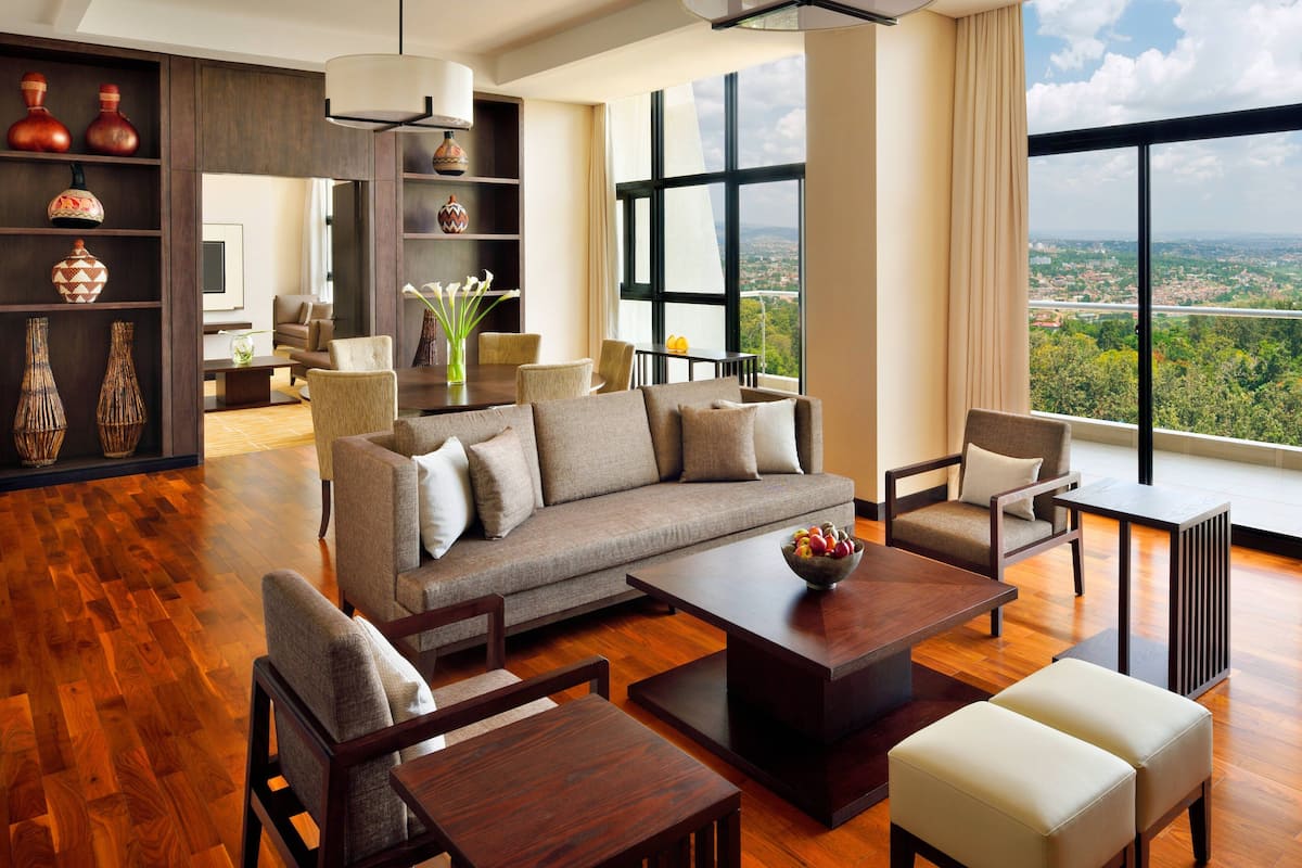 A spacious living room with a large sofa, armchairs, a coffee table, and a dining area. There is a bookshelf with decorative items and a view of a cityscape through large windows.