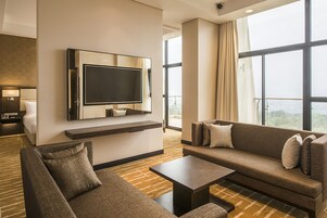 A modern hotel room with a large flat-screen TV mounted on the wall, a sofa, a coffee table, and a view of the city through large windows.