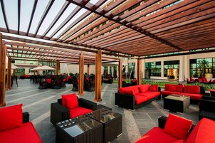 Un área de estar al aire libre con sofás rojos y mobiliario de mimbre bajo una pérgola de madera.