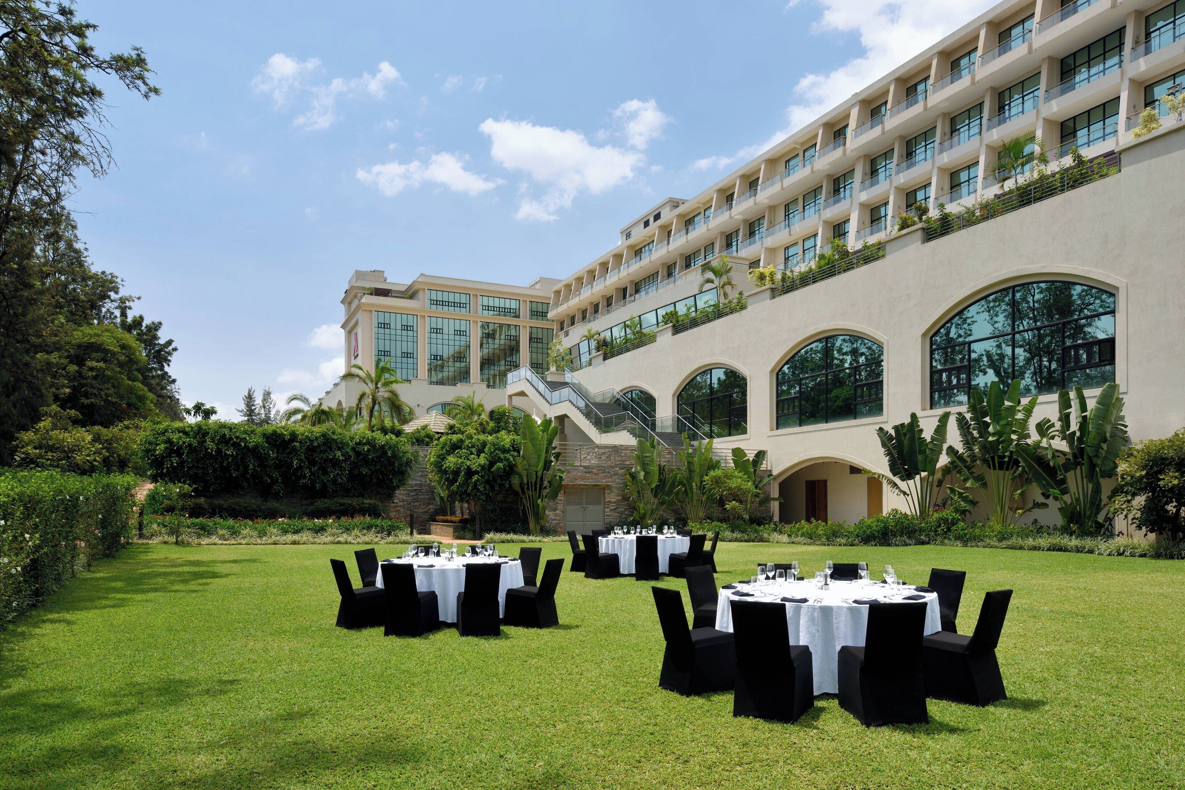 An outdoor dining area with round tables and black chairs, set against a backdrop of a large building with multiple balconies and large windows.