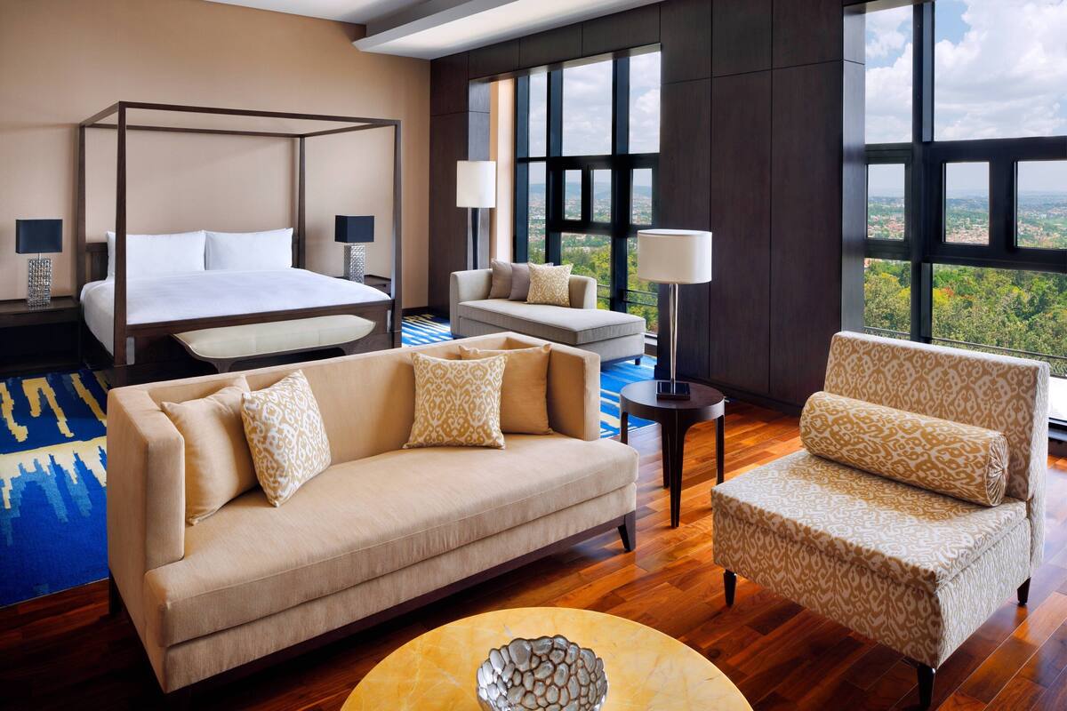A spacious bedroom with a four-poster bed, a sofa, a chair, and a view of the city through large windows.