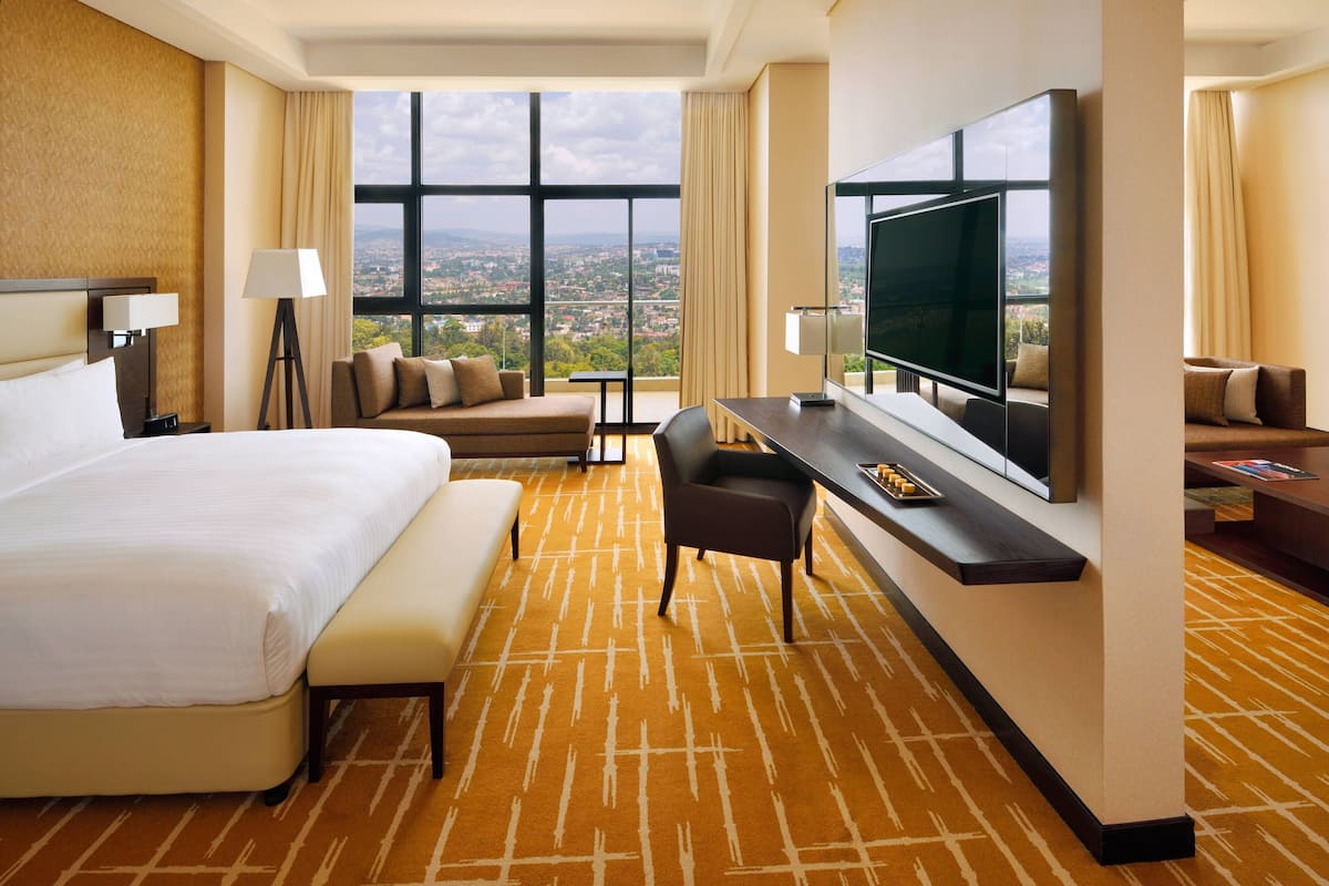 A hotel room with a large bed, a desk with a flat-screen TV, a sofa, and a view of a cityscape through large windows.