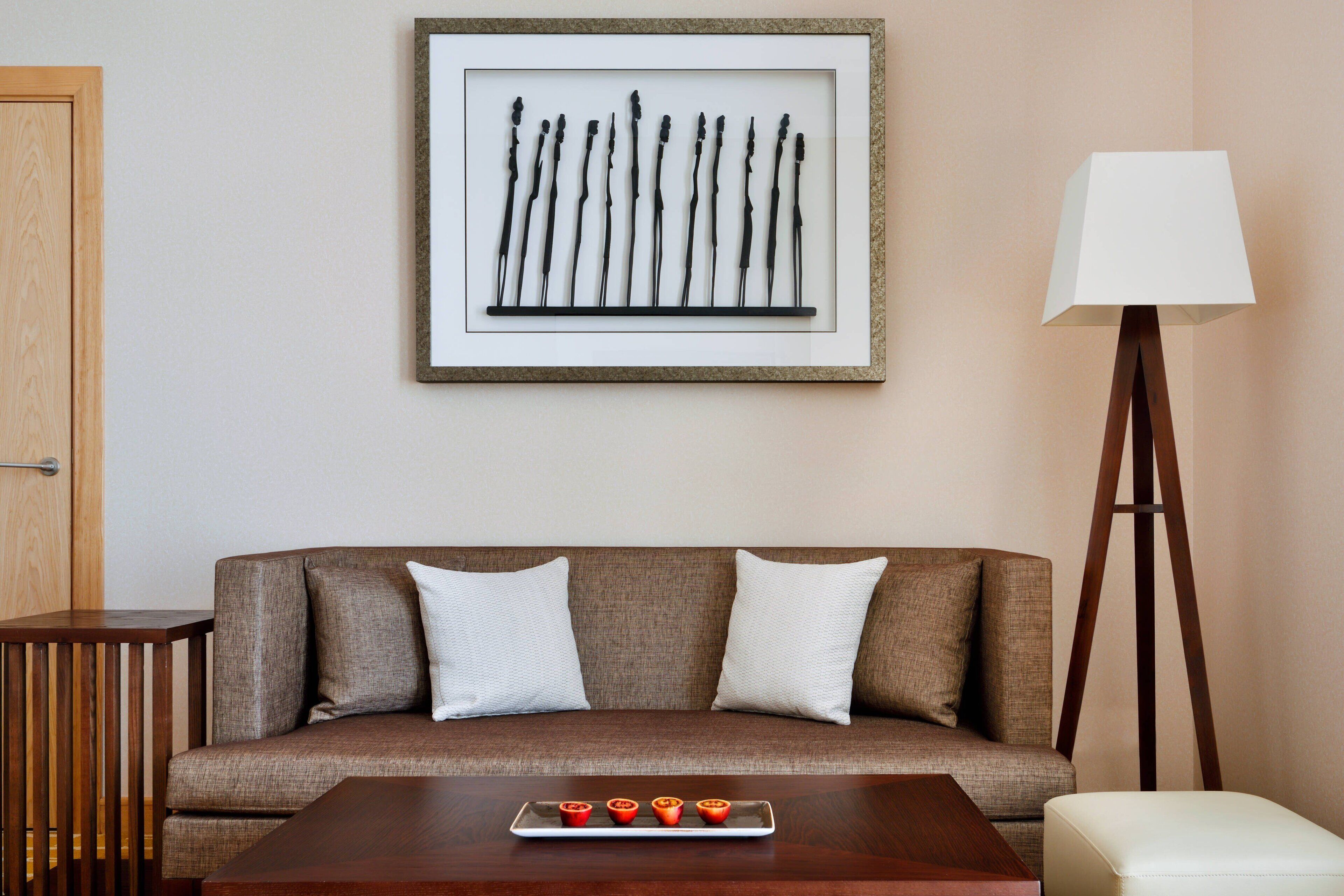 Una habitación con un sofá marrón, dos almohadas blancas, una mesita de madera, una bandeja con objetos rojos, una obra de arte abstracta enmarco y una lámpara de pie.