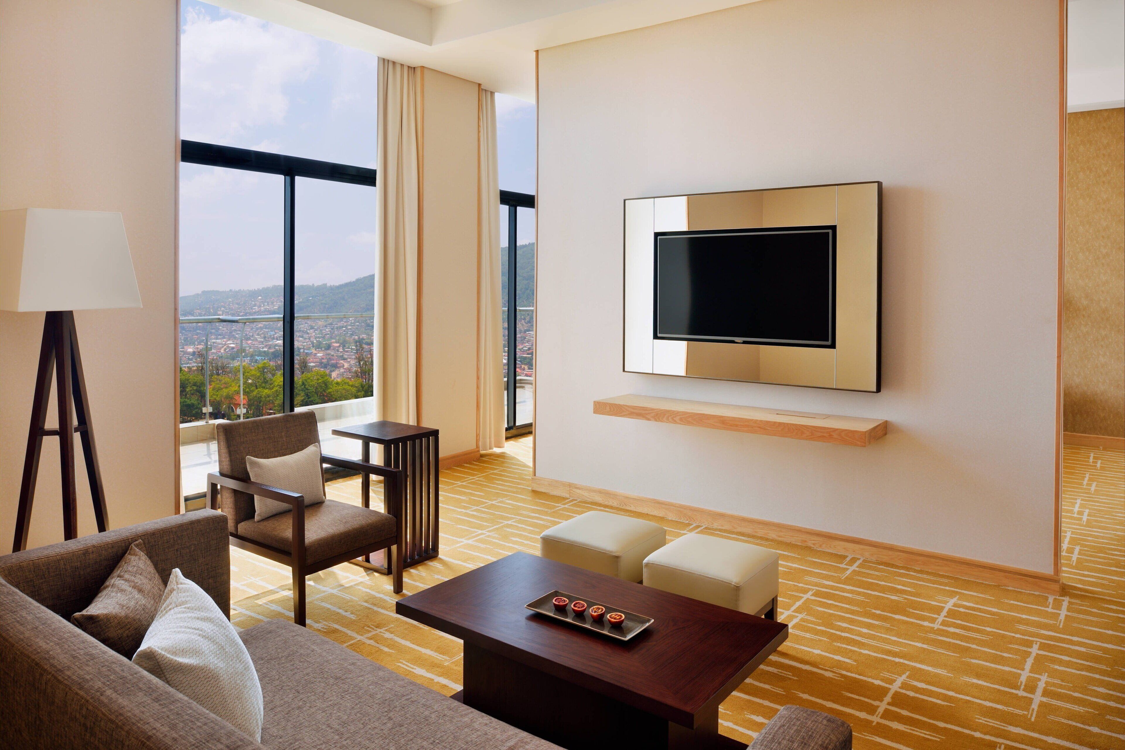 Habitación de hotel con un ventanal grande, un televisor de pantalla plana montado en la pared, un sofá, una mesa de centro y una silla.