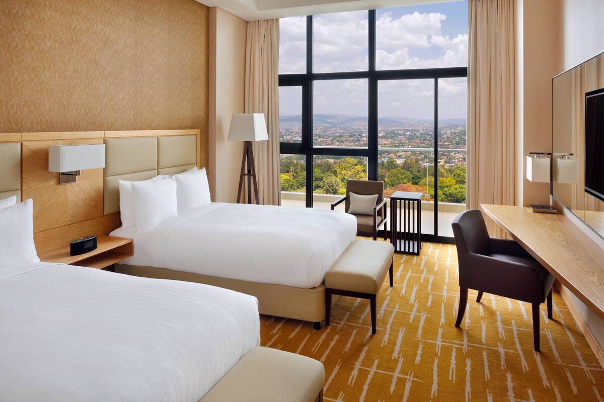 A hotel room with two beds, a desk, a chair, a TV, and a large window offering a city view.