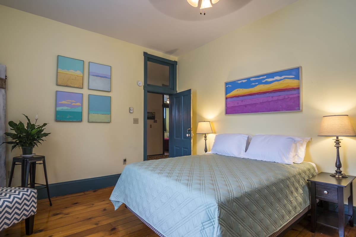A bedroom with a bed, bedside lamps, a nightstand, a chair, and a door leading to another room.