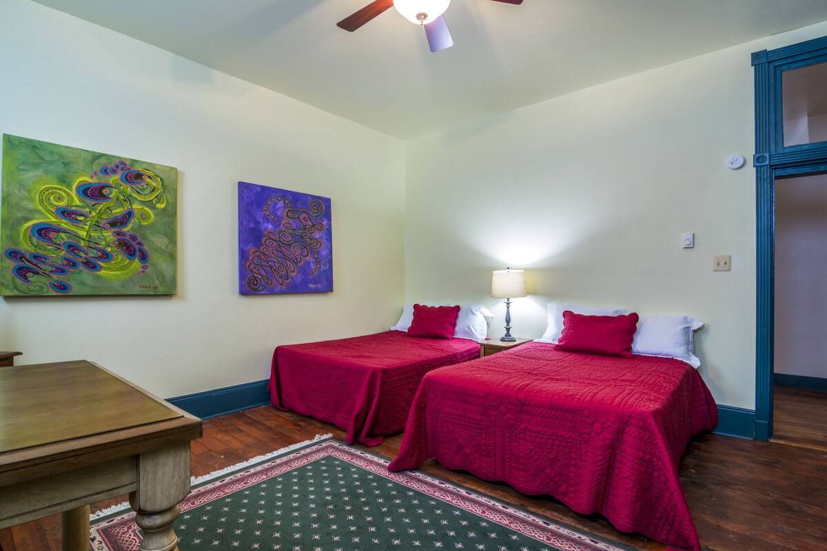 A room with two single beds, a wooden table, a ceiling fan, and two wall-mounted paintings.