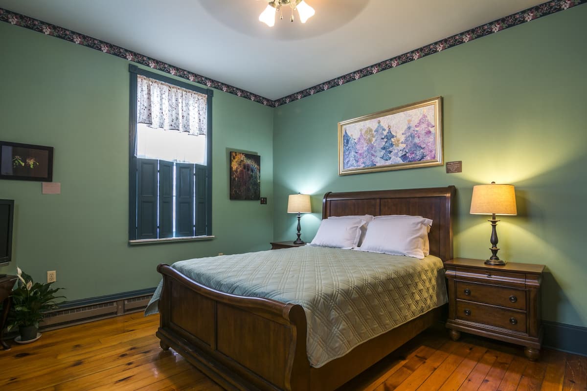 A bedroom with a wooden bed, two bedside tables with lamps, a window with curtains, and a painting on the wall.