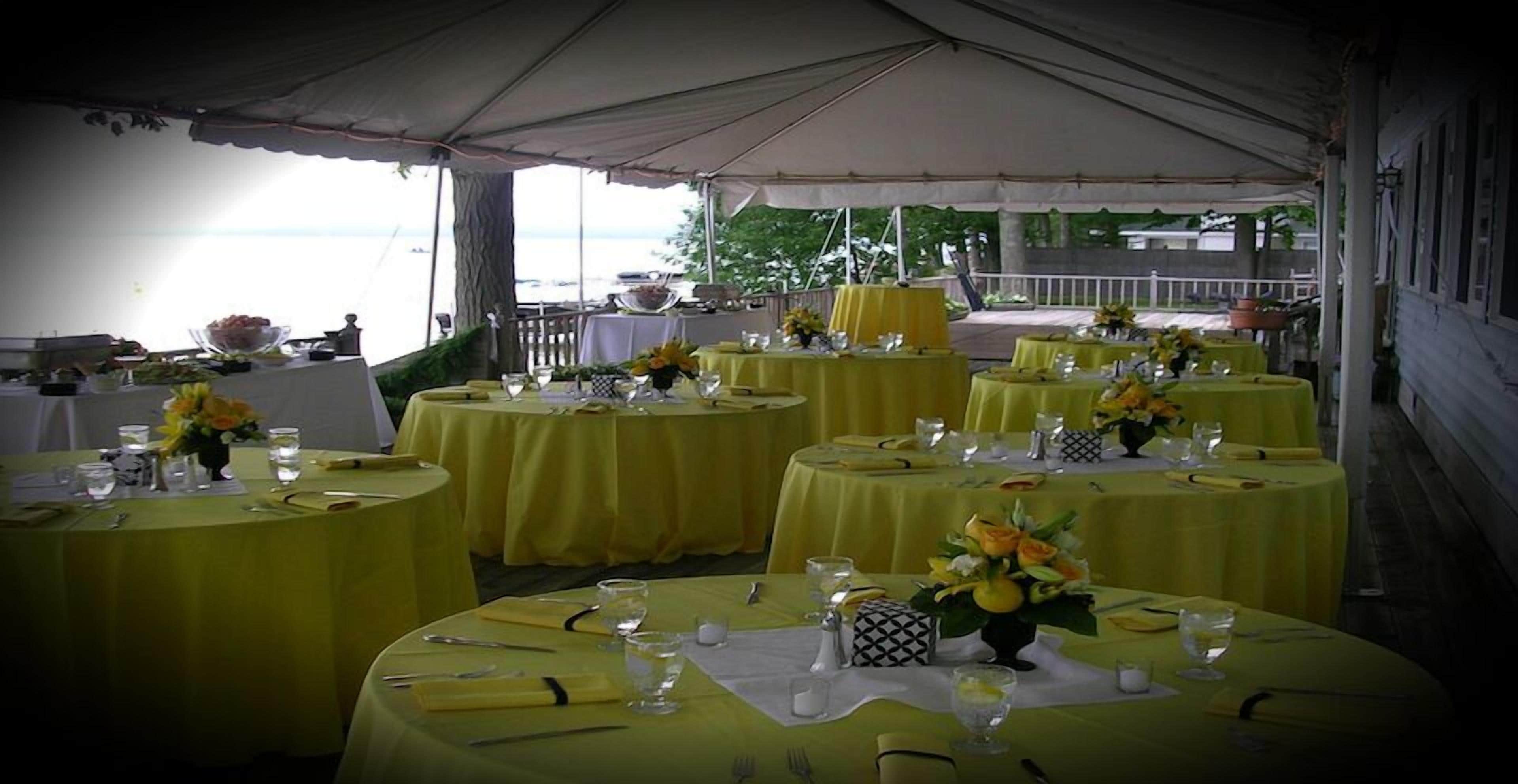 A tented outdoor dining area with round tables covered in yellow tablecloths, each set with cutlery, glasses, and floral centerpieces.