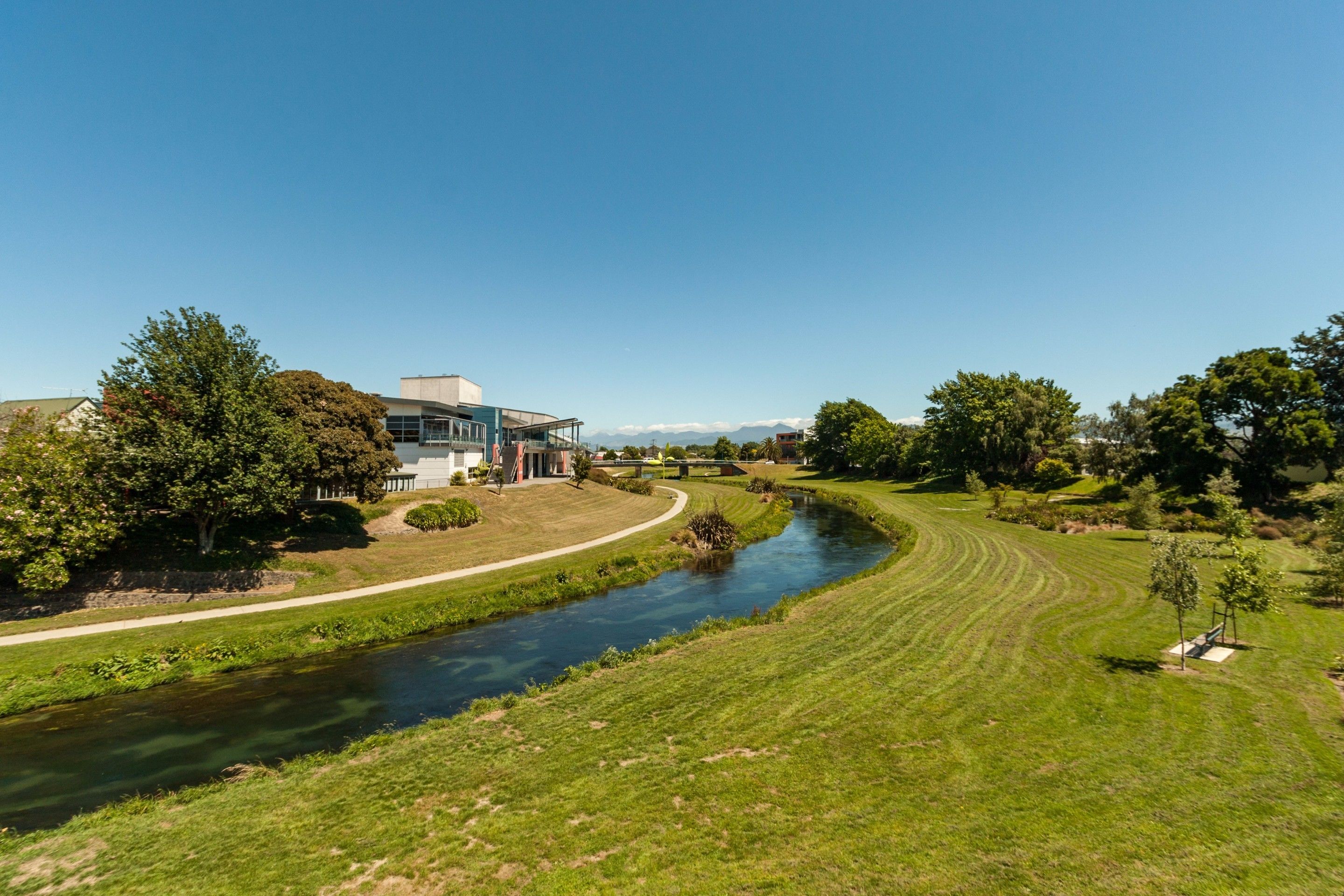 A riverside view with a modern building, grassy banks, and trees.