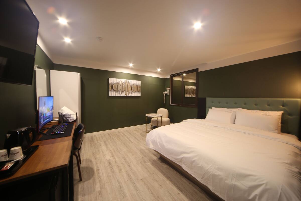 Ein Hotelzimmer mit einem großen Bett, einem Schreibtisch mit Computer, einem Sessel, einem kleinen runden Tisch und einem an der Wand montierten Fernseher.