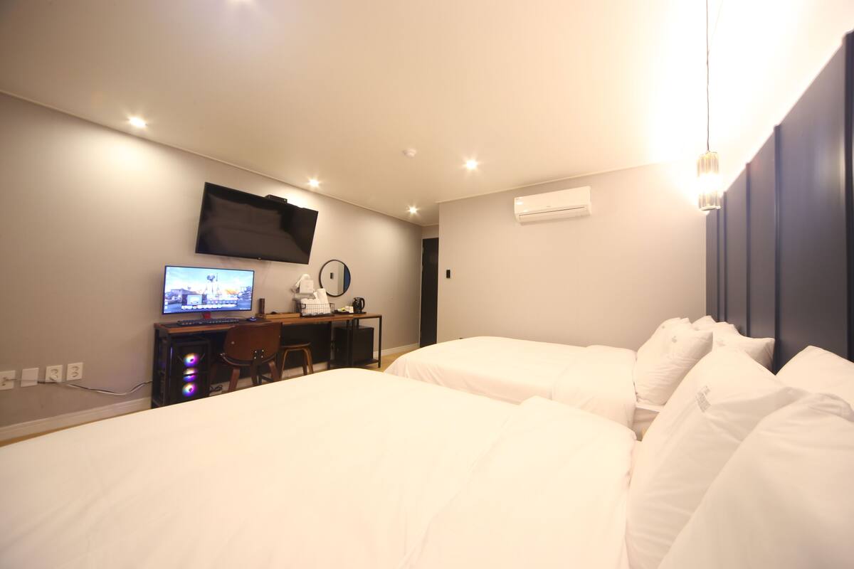 Ein Hotelzimmer mit zwei Betten, einem Schreibtisch mit Computer, einem an der Wand montierten Fernseher und einer Klimaanlage.