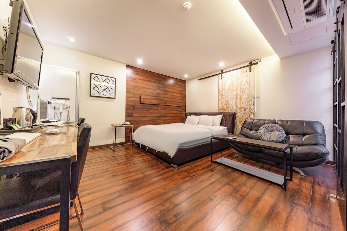 A modern hotel room with a wooden floor, a bed with a white mattress, a leather sofa, a desk with a chair, and a flat-screen TV.