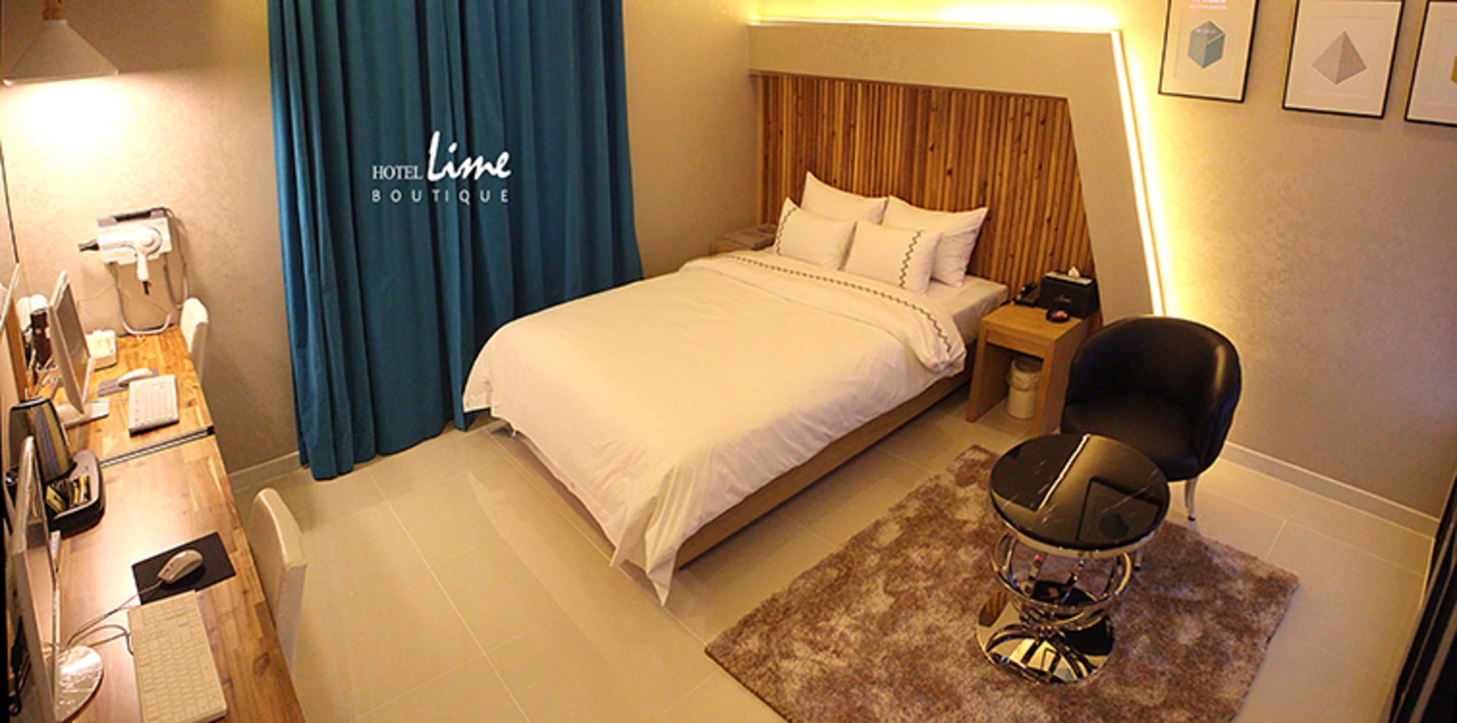 A hotel room with a bed, a desk with a computer, a chair, and a nightstand with a lamp.