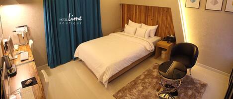 A hotel room with a bed, a desk with a computer, a chair, and a nightstand with a lamp.