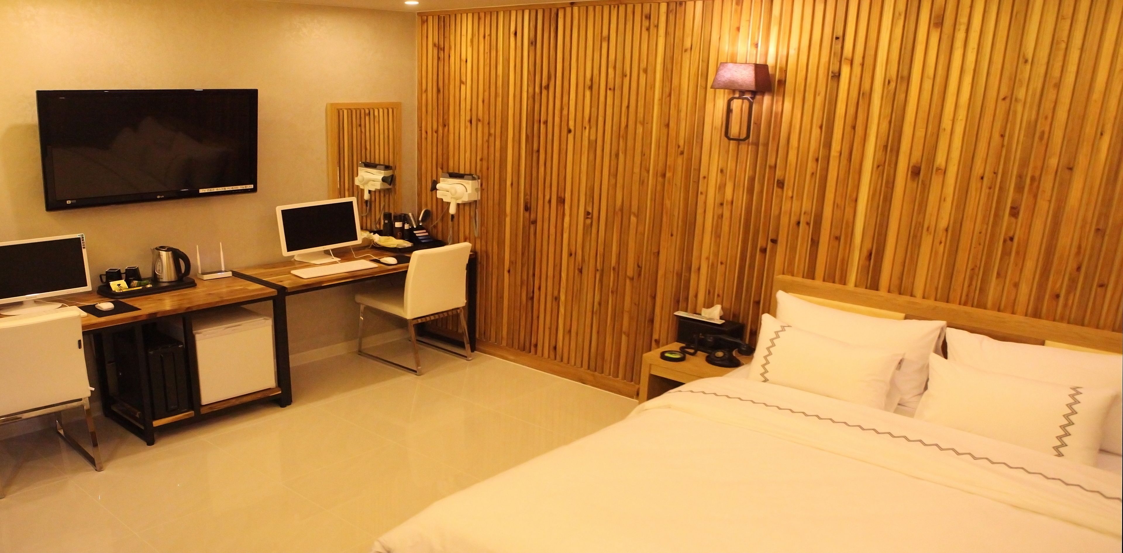 A hotel room with a wooden panel wall, a bed with white linens, a desk with a computer and a chair, a television, and a small bedside table with a lamp.