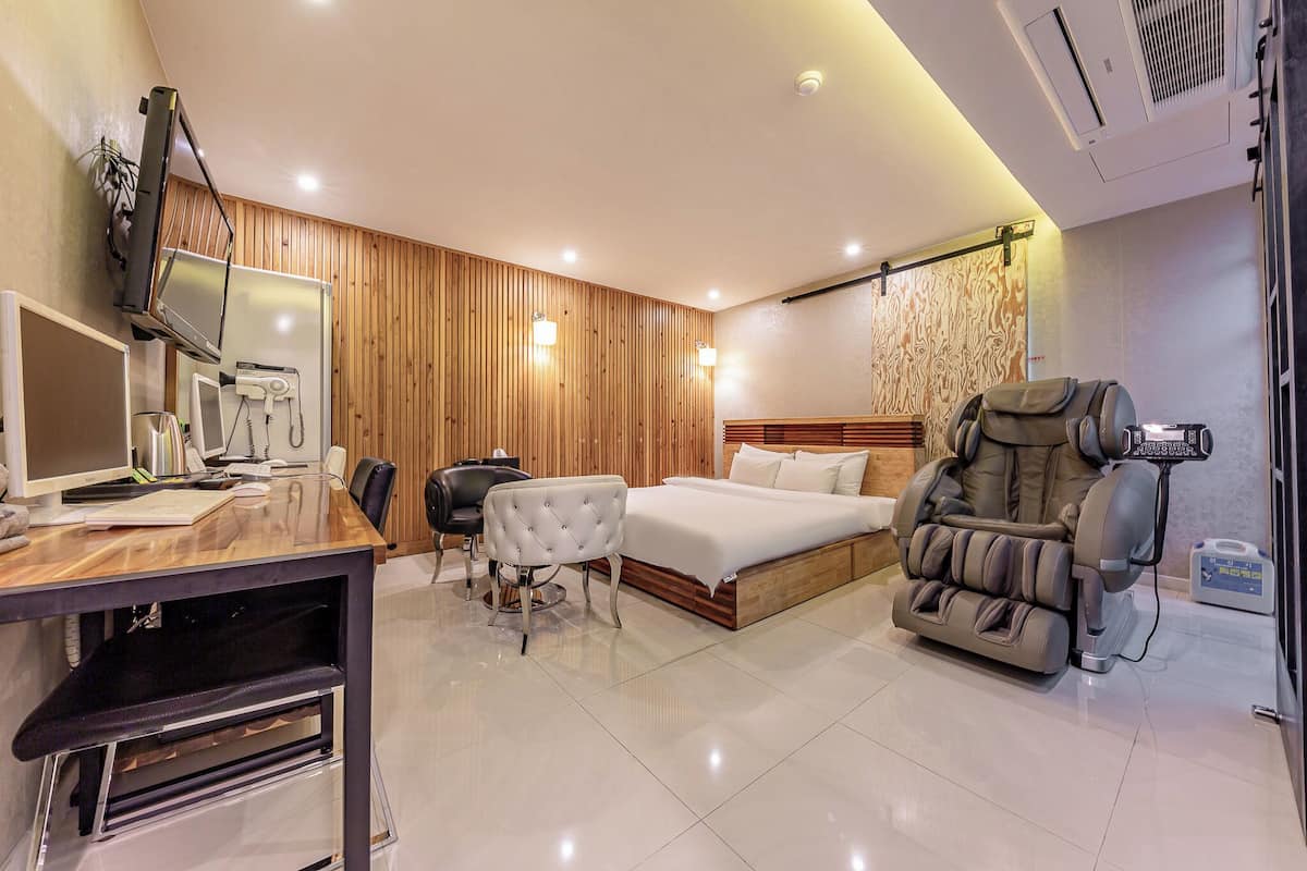 A modern hotel room with a wooden panel wall, a bed, a massage chair, a desk with a computer, and a chair.