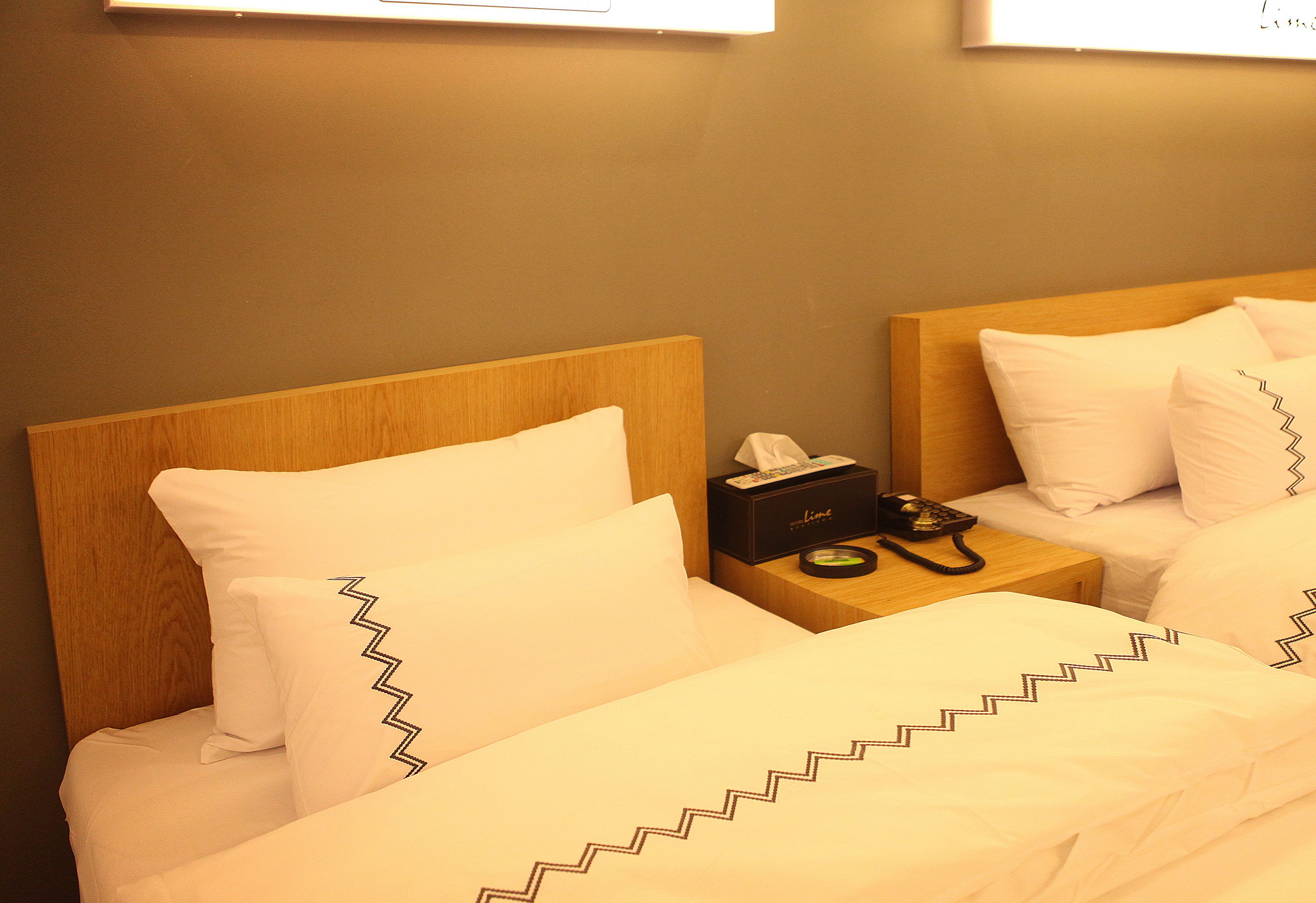 Quarto de hotel com duas camas, cada uma com roupa de cama branca e padrão em zigue-zague. Há uma mesa de cabeceira com uma caixa de lenços e um telefone.