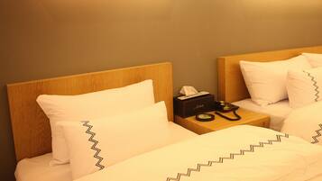 Quarto de hotel com duas camas, cada uma com roupa de cama branca e padrão em zigue-zague. Há uma mesa de cabeceira com uma caixa de lenços e um telefone.