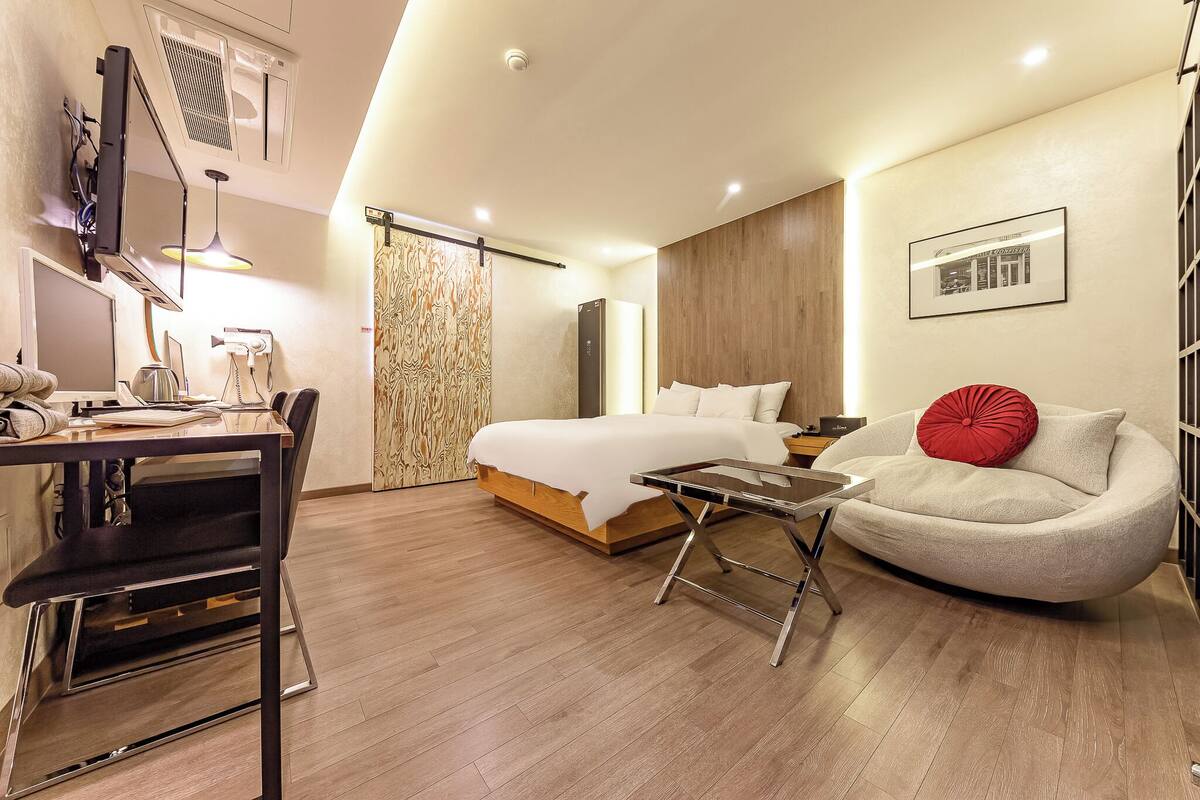 A modern hotel room with a bed, a desk with a chair, a sofa, and a TV.