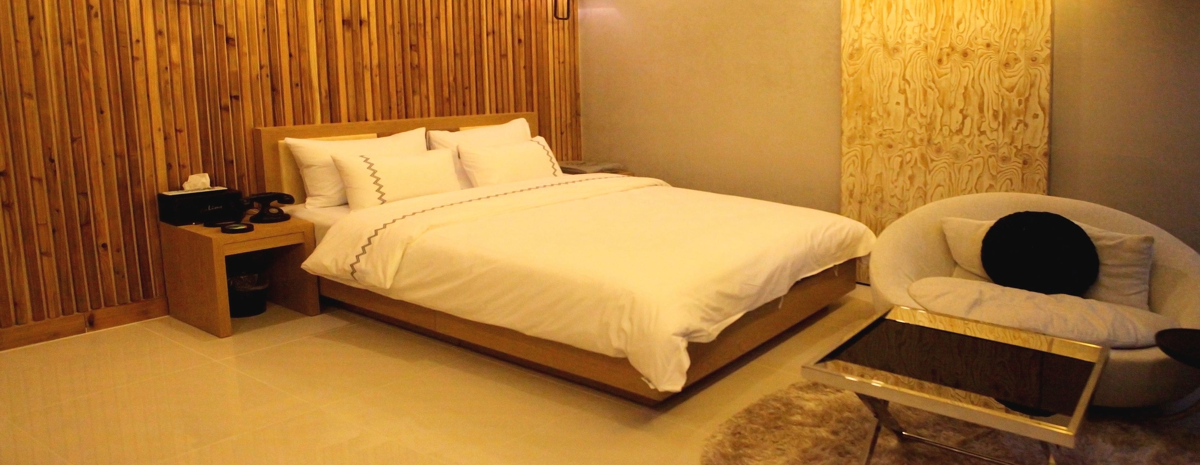 Un lit bien fait, avec du linge de lit blanc et une tête de lit en bois, une table de chevet avec une lampe et une chaise avec un coussin.