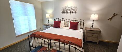 A hotel room with a bed, two bedside tables, a ceiling fan, a window with blinds, and a wall-mounted light fixture.