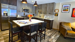 A modern kitchen with a central island, dark wooden cabinets, stainless steel appliances, pendant lighting, and a cozy seating area with a cushioned chair. - Snakedance Condominiums (Taos Ski Valley)