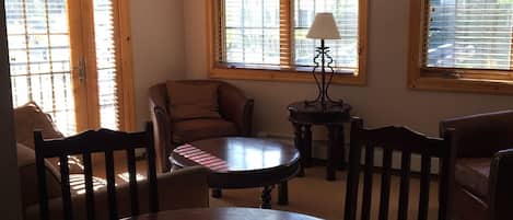 A dining area with a round table, two chairs, a lamp, and a sofa. There are three windows with blinds.